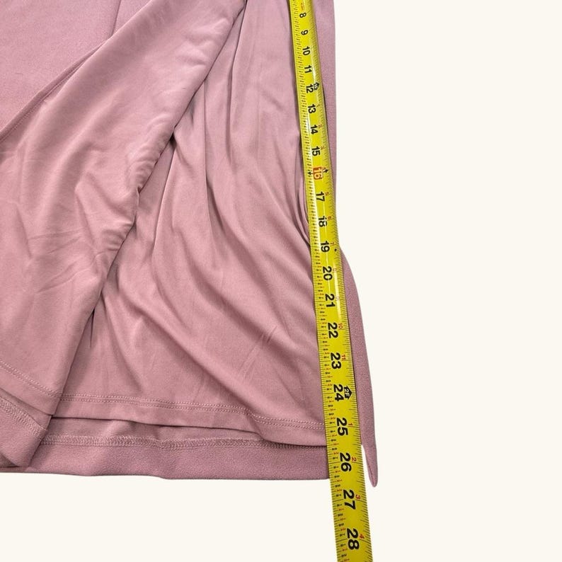 Puede incluir: Una prenda de color rosa polvoriento est&aacute; parcialmente abierta, revelando su interior. Una cinta m&eacute;trica amarilla est&aacute; colocada a lo largo del borde, mostrando medidas de 20 a 71 cm. La tela parece suave y drapeada.