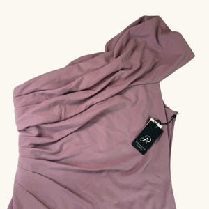 Puede incluir: Un vestido de un solo hombro de color malva con un escote drapeado. El vestido tiene un corpi&ntilde;o ajustado y un dise&ntilde;o asim&eacute;trico. Una etiqueta negra con la marca "Adrianna Papell" est&aacute; adjunta.
