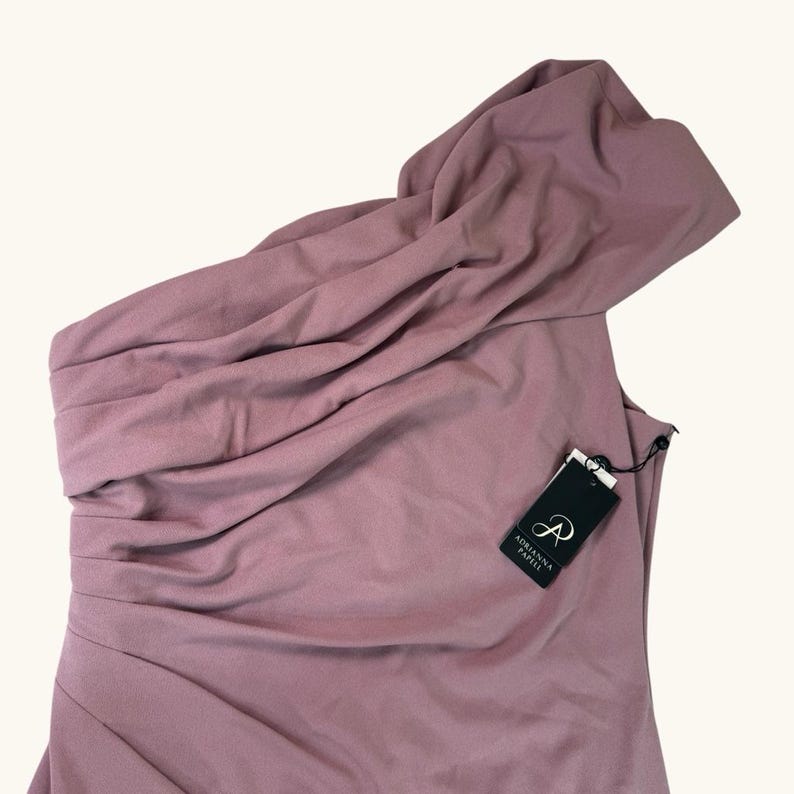 Puede incluir: Un vestido de un solo hombro de color malva con un escote drapeado. El vestido tiene un corpi&ntilde;o ajustado y un dise&ntilde;o asim&eacute;trico. Una etiqueta negra con la marca "Adrianna Papell" est&aacute; adjunta.