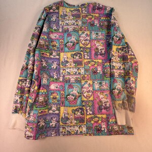 Puede incluir: Una camisa de manga larga con un patrón de cuadrícula colorido y repetitivo de conejos blancos, huevos de Pascua y cestas de flores en tonos morado, azul, amarillo y rosa. La prenda tiene puños acanalados blancos en las mangas y cuello redondo.