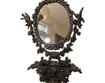 Espejo de tocador antiguo con querubines ornamentados y bandeja para baratijas, con marco floral en bronce dorado.