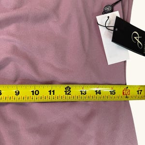 Puede incluir: Primer plano de una tela de color malva con una cinta m&eacute;trica amarilla colocada encima. La cinta mide aproximadamente 40,6 cm. Una etiqueta negra con texto blanco est&aacute; adherida a la tela.