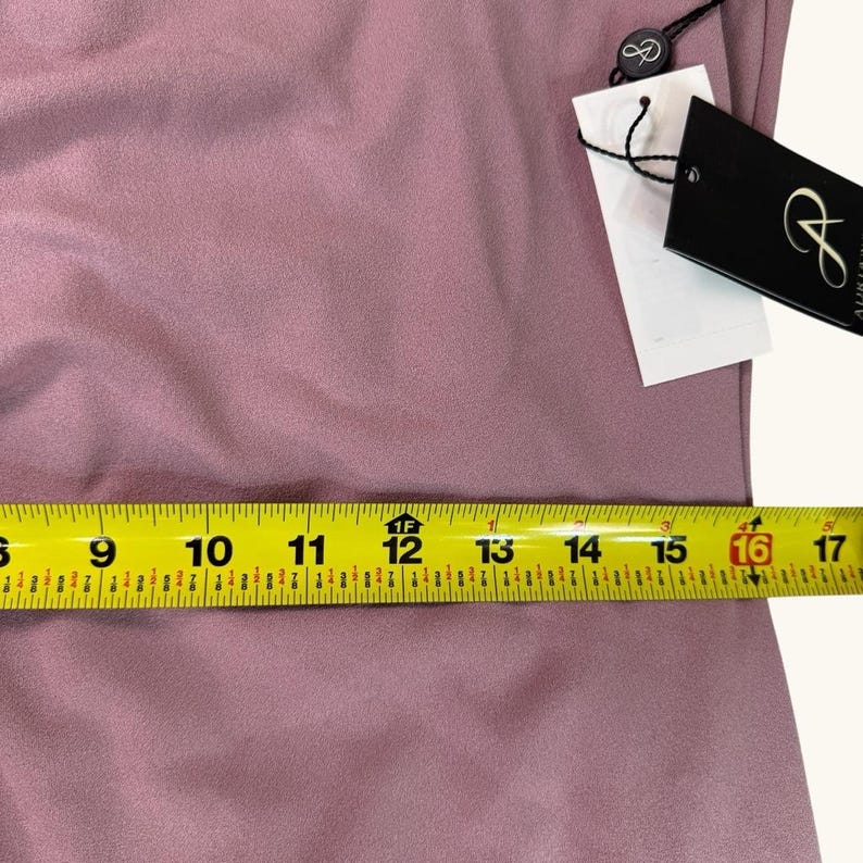 Puede incluir: Primer plano de una tela de color malva con una cinta m&eacute;trica amarilla colocada encima. La cinta mide aproximadamente 40,6 cm. Una etiqueta negra con texto blanco est&aacute; adherida a la tela.