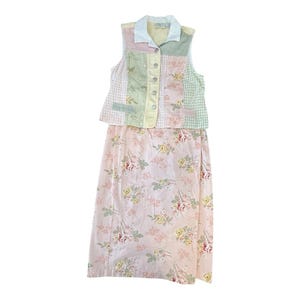 May include: A two-piece outfit featuring a patchwork vest and a floral print skirt. The vest has a white collar and button closure, with pastel-colored patches. The skirt is a light pink with a floral pattern.