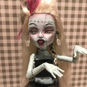 May include: A doll with a pale blue complexion, styled after a monster. The doll has blonde hair with pink streaks, stitched features, and a black top. The doll is wearing a black skirt and a silver belt. The doll has a unique, stylized appearance.