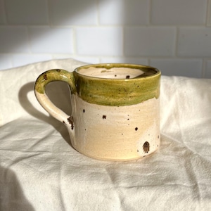May include: Handmade ceramic mug with a cream base and a green rim. The mug has a rounded handle and is speckled with brown spots. The mug is on a white cloth.