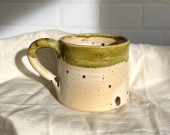 May include: Handmade ceramic mug with a cream-colored base and a green rim. The mug has a rounded handle and is speckled with brown spots. The mug is sitting on a white cloth.