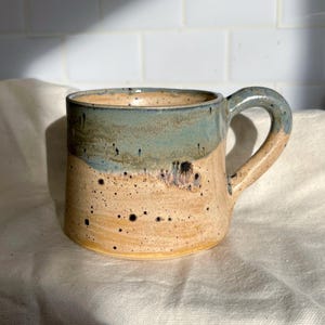 May include: Handmade ceramic mug with a speckled tan base and a blue-green upper section. The mug features a rounded handle and is adorned with dark speckles, showcasing a rustic, artisanal aesthetic. A unique piece for enjoying beverages.