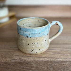 May include: A ceramic mug with a light blue rim and handle, and a beige body speckled with black dots. The mug has a curved handle and a slightly textured surface. Ideal for tea or coffee.