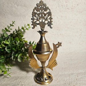 Pode incluir: Um frasco de perfume de latão vintage com uma rolha ornamentada em forma de árvore. O frasco tem um corpo bulboso e é suportado por uma base decorativa com detalhes em forma de asa. O design geral é detalhado e antigo.
