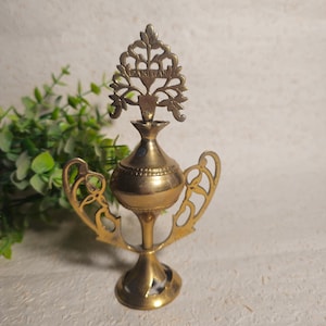 Puede incluir: Un quemador de incienso de latón vintage con una parte superior decorativa en forma de árbol que dice "PAKISTAN". El quemador tiene un cuerpo bulboso, asas ornamentadas y una base de pedestal. El diseño general es intrincado y detallado.