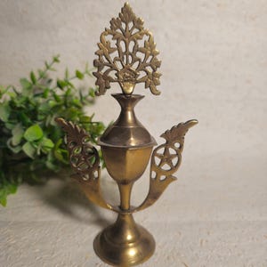 Pode incluir: Um aplicador de kohl de latão vintage. O aplicador apresenta um design decorativo em forma de árvore no topo, um corpo hexagonal e asas laterais ornamentadas com recortes em forma de estrela. A base é redonda e alargada.