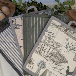 May include: A collection of four quilted pot holders with gray trim and hanging loops. Designs include a floral pattern, vertical stripes, and a farm-themed print with a basket of produce, a rooster, and a windmill. The pot holders are arranged on a white surface.