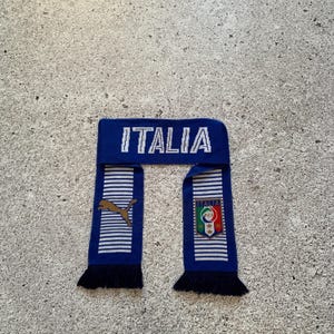 May include: A blue and white striped scarf with the word "ITALIA" in white letters. The scarf has a Puma logo and an Italian flag emblem. The scarf has black fringe.