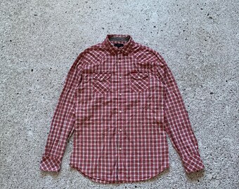 May include: A long-sleeved, button-down shirt in a red and white checkered pattern. The shirt features two chest pockets with decorative stitching and a classic collar. The cuffs are buttoned, and the shirt is laid flat on a textured surface.