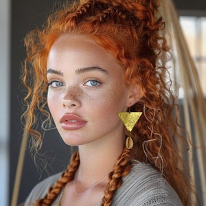 Puede incluir: Una persona con cabello rizado rojo peinado en un moño alto y trenzas, con pendientes triangulares dorados con un colgante de concha. La persona tiene ojos azules y pecas.