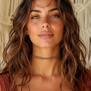 May include: A close-up portrait of a person wearing a beaded choker necklace. The necklace features two strands of small, colorful beads in shades of orange, brown, and gold. The person has long, wavy brown hair and is wearing a rust-colored top.