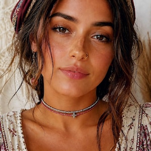 May include: A woman wearing a burgundy headband and a floral print top. A layered choker necklace with a small cross pendant is around her neck. The necklace has red and silver beads.