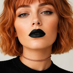 Può includere: Un primo piano di una persona con capelli rossi corti e mossi e rossetto nero. La persona indossa un top nero e una delicata collana girocollo con perline color rame. Gli occhi sono accentuati con eyeliner nero.