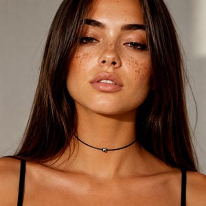 Può includere: Primo piano di una persona che indossa una collana girocollo nera con una piccola perla color argento. La persona ha i capelli castano scuro e lentiggini. Lo sfondo è grigio neutro.