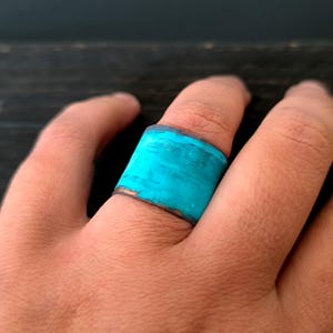 Può includere: Un anello largo con una fascia turchese blu e un bordo in metallo scuro. L'anello è indossato su un dito.