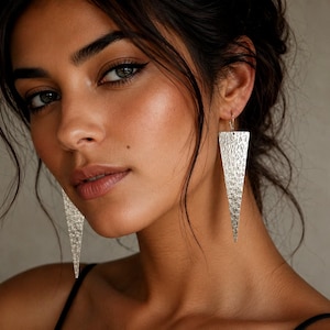 Può includere: Un paio di lunghi orecchini pendenti triangolari realizzati in metallo color argento. Ogni orecchino presenta una superficie martellata e testurizzata che cattura la luce, sospeso a un semplice gancio che attraversa il lobo dell'orecchio.