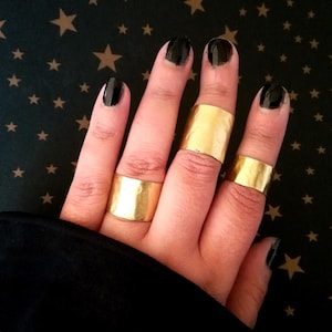 Puede incluir: Una mano con cuatro anillos de oro. Los anillos son anchos y planos, con una textura martillada. La mano está sobre un fondo negro con estrellas doradas.