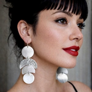 Può includere: Orecchini pendenti in argento con un design a strati. Gli orecchini presentano una serie di forme circolari e semicircolari, creando un look moderno e geometrico. La persona indossa capelli scuri e rossetto rosso.