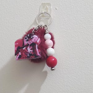 May include: A keychain with a red and black anime character charm, a fluffy burgundy pom-pom, and a beaded accent of white, pink, and red spheres. The keychain is attached to a silver keyring.