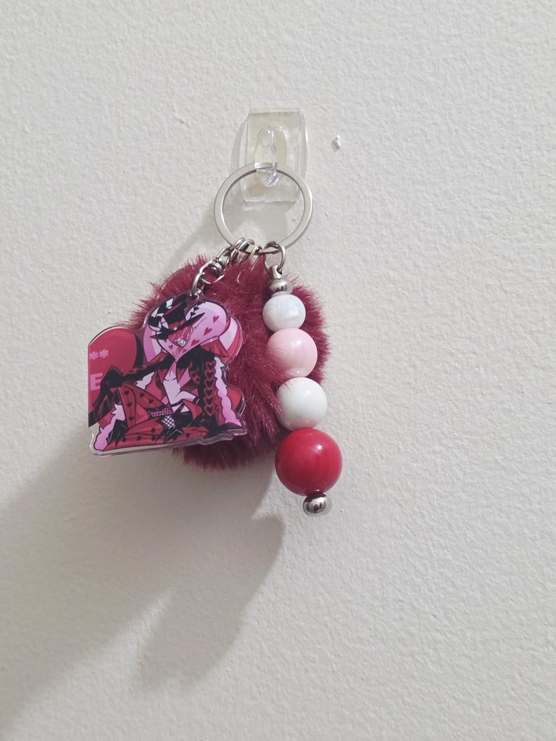 May include: A keychain with a red and black anime character charm, a fluffy burgundy pom-pom, and a beaded accent of white, pink, and red spheres. The keychain is attached to a silver keyring.