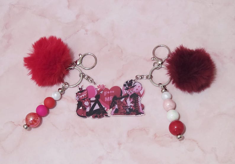 May include: Two keychains with silver rings, red pom-poms, and beaded accents. One keychain has a red, pink, and white bead strand, while the other has a white, pink, and red bead strand. Both have a heart-shaped charm with the text "F*** ME".