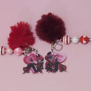 May include: Two keychains with red and white beaded accents and fluffy burgundy pom-poms. Each keychain has a heart-shaped charm with a cartoon character and text that reads "Love Your Self" and "Miss You".