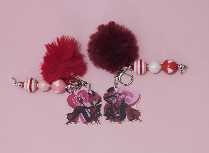 May include: Two keychains with red and white beaded accents and fluffy burgundy pom-poms. Each keychain has a heart-shaped charm with a cartoon character and text that reads "Love Your Self" and "Miss You".