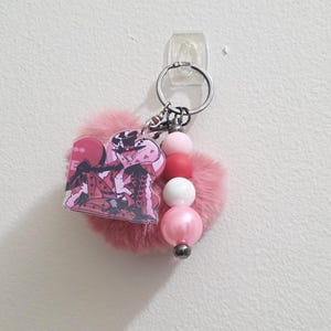 May include: A pink keychain with a fluffy heart-shaped charm, a cartoon character, and a string of pink and white beads. The keychain has a silver ring for attaching keys.