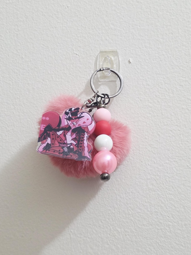 May include: A pink keychain with a fluffy heart-shaped charm, a cartoon character, and a string of pink and white beads. The keychain has a silver ring for attaching keys.