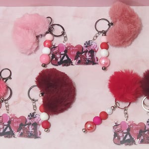 May include: Keychains with fluffy heart-shaped accents in shades of pink and red. Each keychain features a beaded strand with pink, white, and red beads, and a charm with a graphic design and the text "F**k ME".