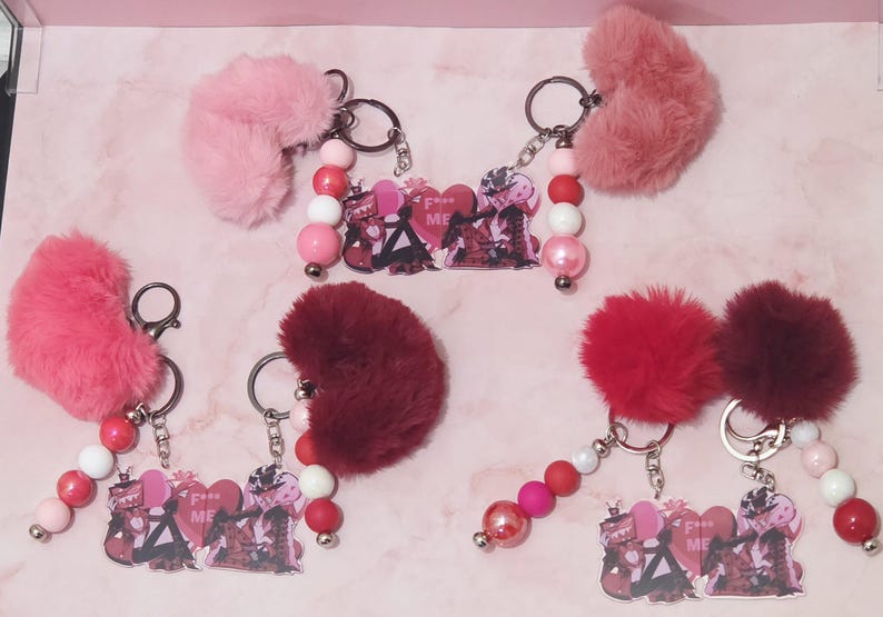 May include: Keychains with fluffy heart-shaped accents in shades of pink and red. Each keychain features a beaded strand with pink, white, and red beads, and a charm with a graphic design and the text "F**k ME".