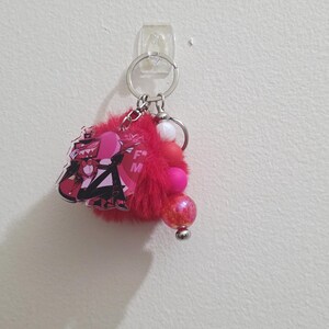 May include: A red keychain with a cartoon character design, a fluffy pom-pom, and decorative beads. The keychain features a silver ring and clasp for attaching keys or accessories. The design includes pink and black colors.