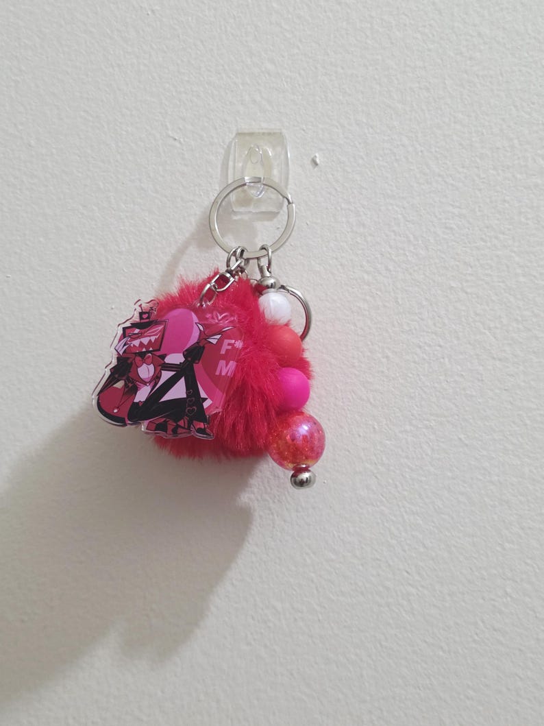 May include: A red keychain with a cartoon character design, a fluffy pom-pom, and decorative beads. The keychain features a silver ring and clasp for attaching keys or accessories. The design includes pink and black colors.
