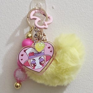 May include: A pink and yellow keychain with a heart-shaped charm featuring a cartoon character. The charm is adorned with a yellow heart-shaped gem and a fluffy yellow pom-pom. Additional pink and gold beads dangle from the keychain.