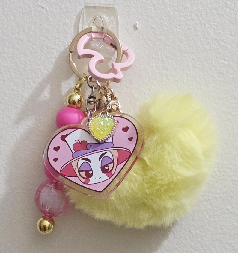 May include: A pink and yellow keychain with a heart-shaped charm featuring a cartoon character. The charm is adorned with a yellow heart-shaped gem and a fluffy yellow pom-pom. Additional pink and gold beads dangle from the keychain.
