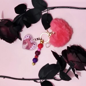 May include: A pink keychain with a fluffy heart, a heart-shaped charm with a cartoon character, and a beaded charm. The keychain is accented with gold-colored hardware. Black roses are arranged on a pink background.