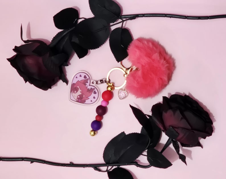 May include: A pink keychain with a fluffy heart, a heart-shaped charm with a cartoon character, and a beaded charm. The keychain is accented with gold-colored hardware. Black roses are arranged on a pink background.