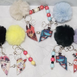 May include: Keyrings with cartoon character designs. Each keyring features a fluffy pom-pom in various colours, such as black, yellow, and blue. The keyrings have heart-shaped charms with cartoon characters and beaded accents.