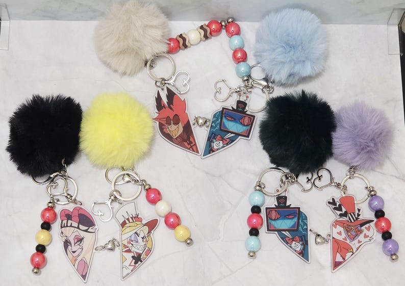 May include: Keyrings with cartoon character designs. Each keyring features a fluffy pom-pom in various colours, such as black, yellow, and blue. The keyrings have heart-shaped charms with cartoon characters and beaded accents.