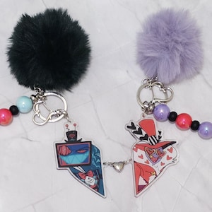 May include: Two keyrings with cartoon character designs. One keyring has a dark green fluffy pom-pom, the other a light purple pom-pom. Each keyring has a beaded strand with blue, pink, and black beads. The charms are connected with a small heart.