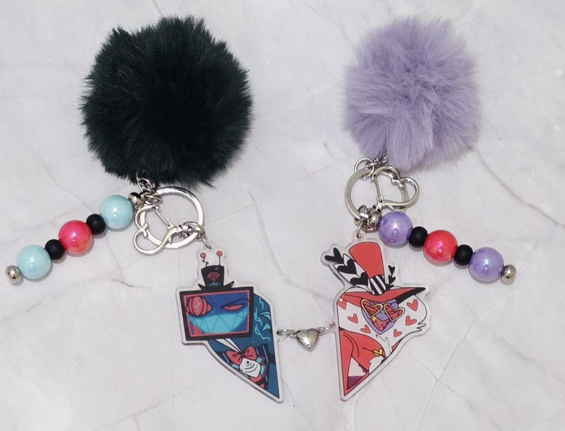 May include: Two keyrings with cartoon character designs. One keyring has a dark green fluffy pom-pom, the other a light purple pom-pom. Each keyring has a beaded strand with blue, pink, and black beads. The charms are connected with a small heart.