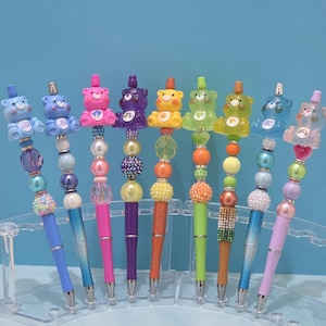 May include: A collection of colorful pens with decorative bear toppers. Each pen features a different color bear, including blue, pink, yellow, and green. The pens have beaded accents and are displayed in a clear holder. The pens are a fun and unique stationery item.