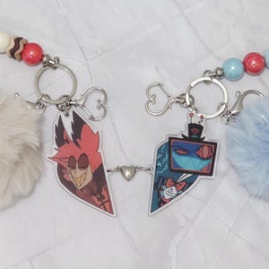 May include: Two keyrings with cartoon character pendants, fluffy pom-poms, and beaded accents. One keyring has a red and brown bead strand, a cream pom-pom, and a red cartoon character. The other has a blue pom-pom and a blue and red bead strand.