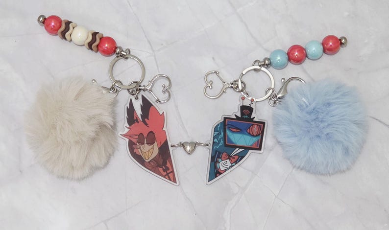 May include: Two keyrings with cartoon character pendants, fluffy pom-poms, and beaded accents. One keyring has a red and brown bead strand, a cream pom-pom, and a red cartoon character. The other has a blue pom-pom and a blue and red bead strand.